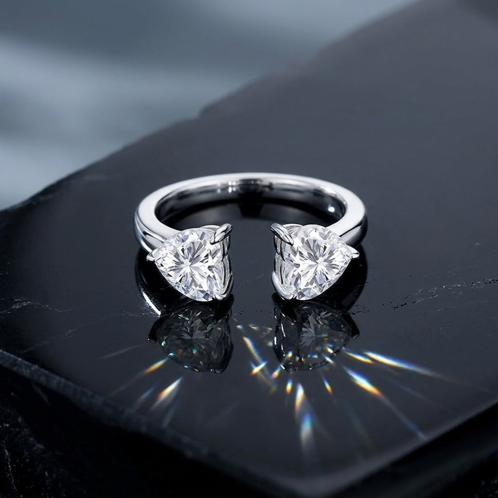 
Sterling silver open adjustable cuff ring featuring dual heart-shaped prong-set cubic Moissanite gemstones resting on a dark reflective surface with prismatic light flares by J.Collins
