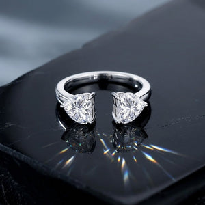 
Sterling silver open adjustable cuff ring featuring dual heart-shaped prong-set cubic Moissanite gemstones resting on a dark reflective surface with prismatic light flares by J.Collins

