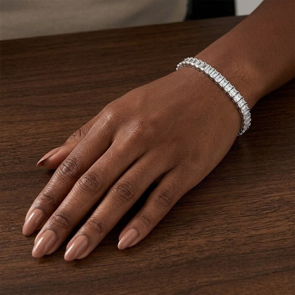 Classic single-row sterling silver tennis bracelet set with emerald-cut cubic Moissanite crystals worn on wrist resting on dark wood table by model Tiffany by J.Collins
