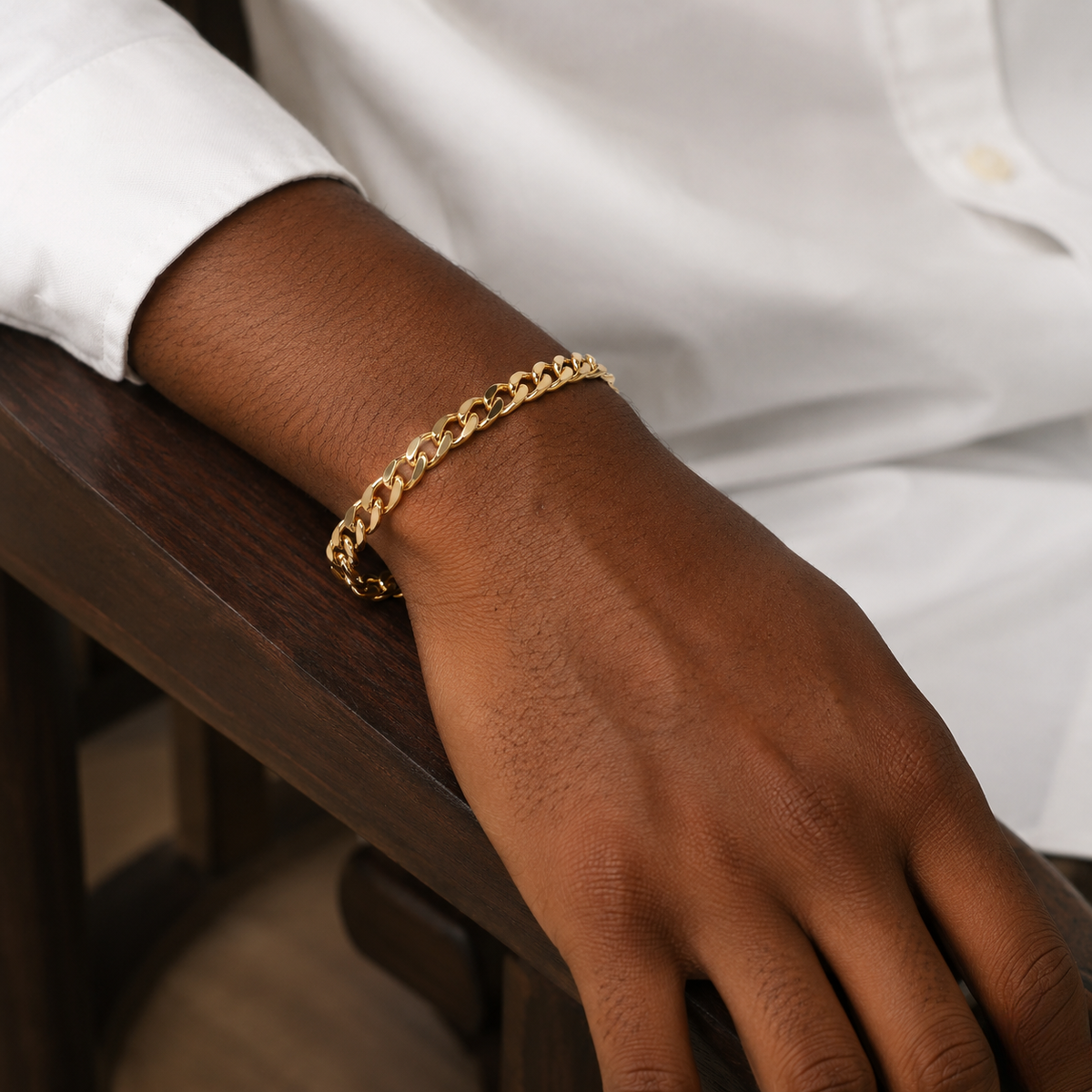 Close up of man's wrist in white shirt resting on a wood chair, wearing gold figaro curb chain bracelet, luxury men's accessory - J.Collin Jewelry