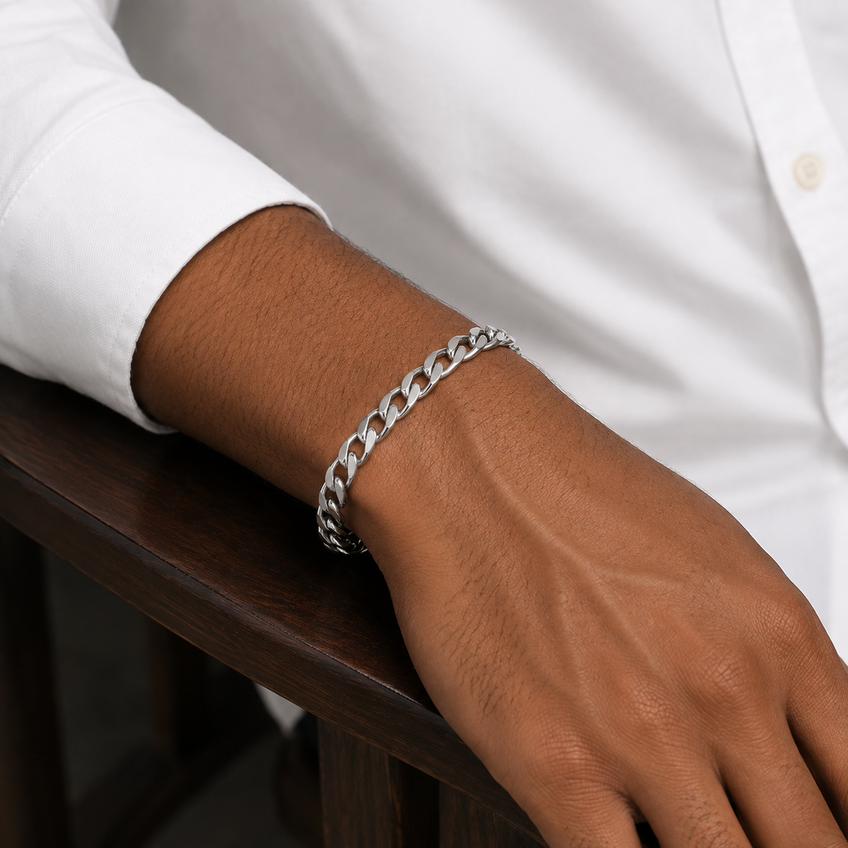 Male model wearing white dress shirt with rolled sleeve, silver thick 5mm Figaro chain bracelet on wrist, wood chair background, men's stainless steel - J.Collins Jewelry