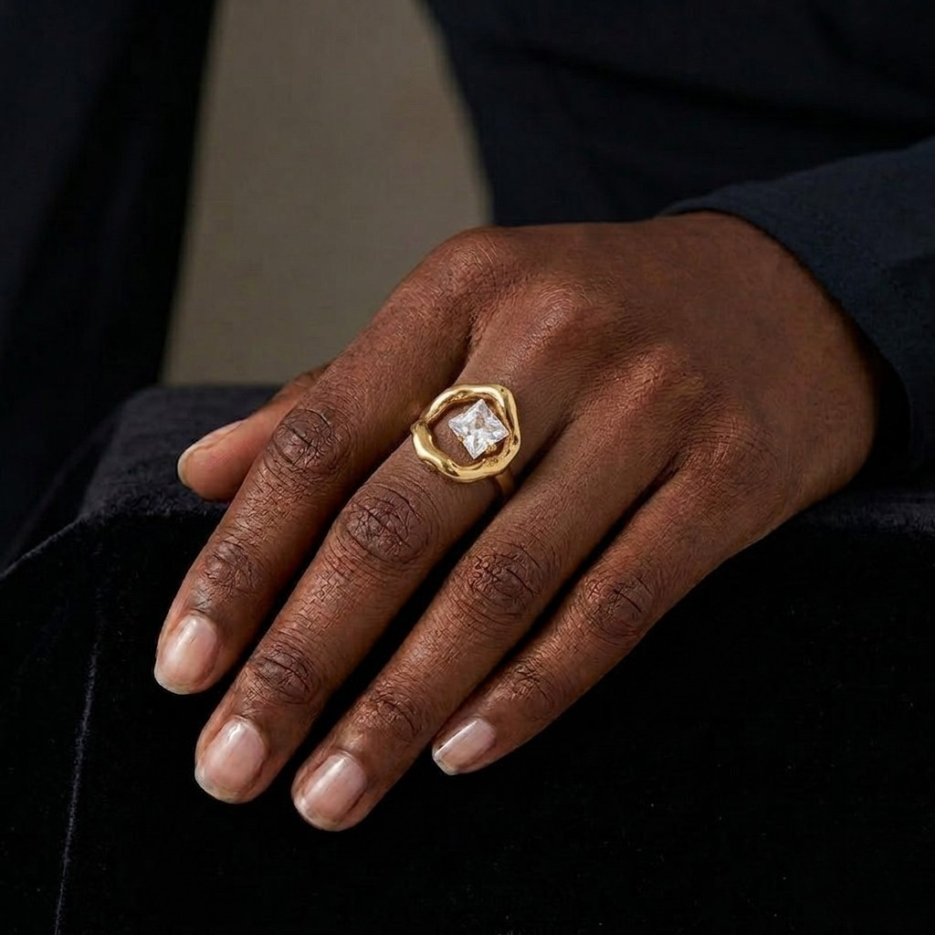 A close-up shot of a hand resting against a black velvet surface, showcasing a statement gold ring worn on the middle finger. The ring features an open, organic-shaped molten gold frame encasing a sparkling, faceted square-cut clear gemstone tilted at the center. The hand is styled with natural manicured nails and wears a long-sleeved black garment against a soft, dark background.
