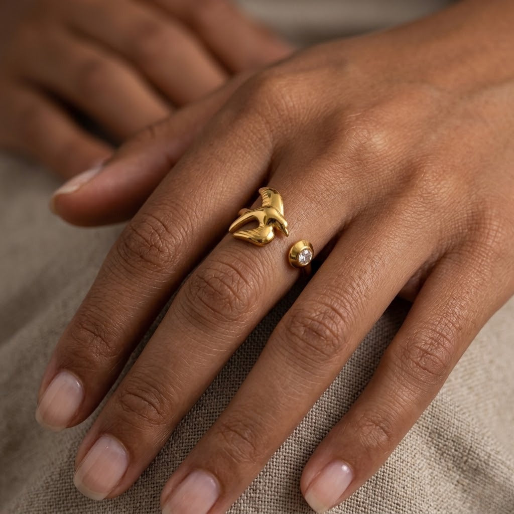 A close-up of a hand wearing two gold rings: a larger sculptural ring with a small central clear stone and a smaller matching ring with a single round clear stone, set against a neutral beige fabric background - J.Collins Jewelry