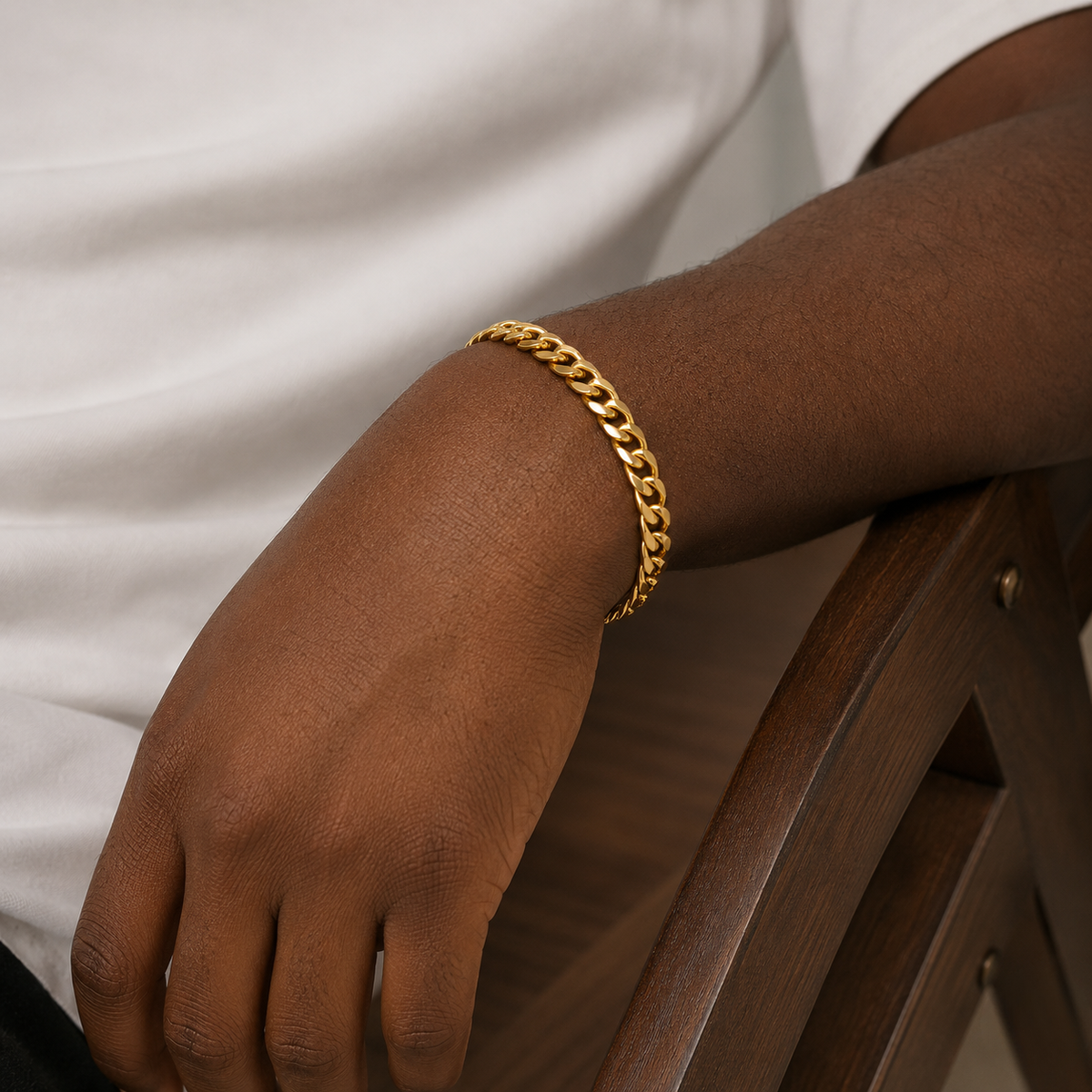 A close-up of a person's forearm and hand wearing a thick gold chain bracelet, resting on a dark wooden chair arm while wearing a white t-shirt - J.Collins Jewelry