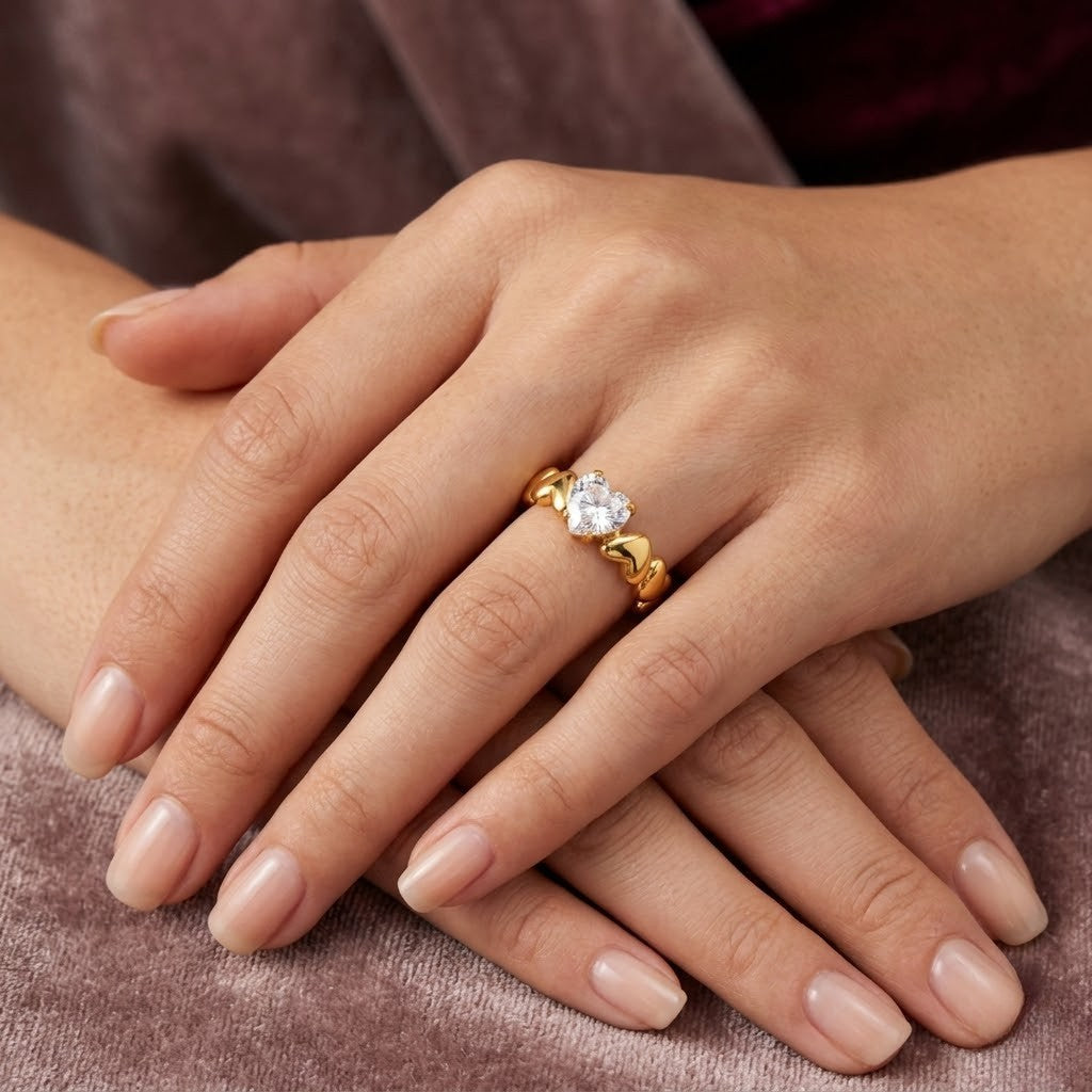 Gold heart solitaire ring with a clear gemstone on a woman's hand resting on fabric - J.Collins Jewelry