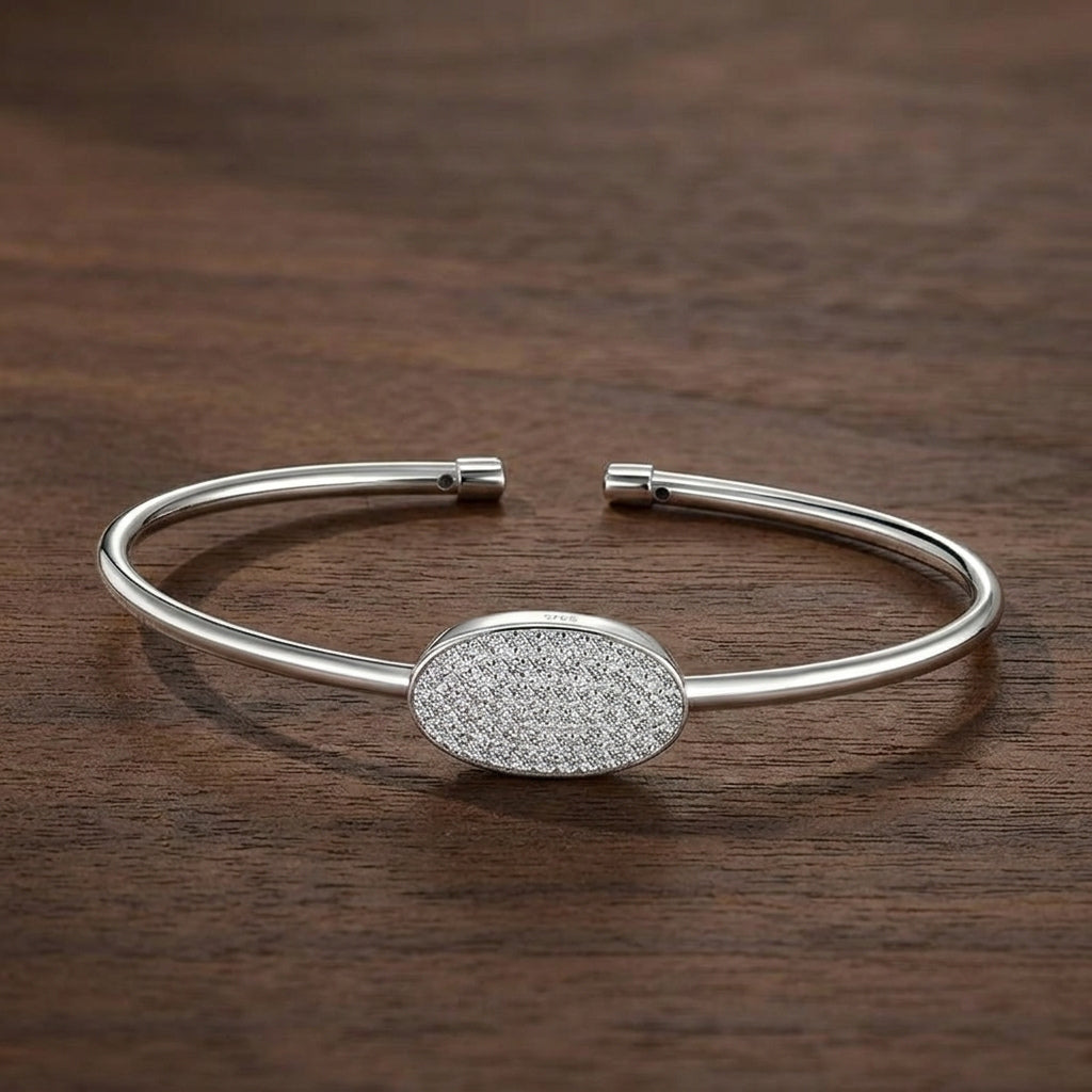 A polished silver cuff bracelet with a central oval pavé-set stone cluster, displayed on a dark brown wooden surface.