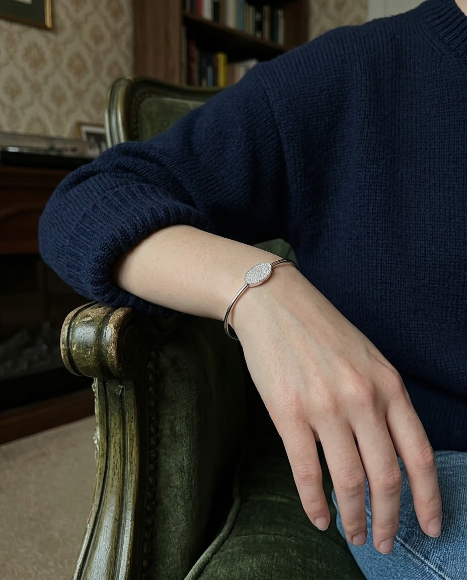 A person wearing a dark navy blue knit sweater and light blue jeans rests their arm on an antique green armchair, wearing a delicate rose gold bangle bracelet with a round white stone.