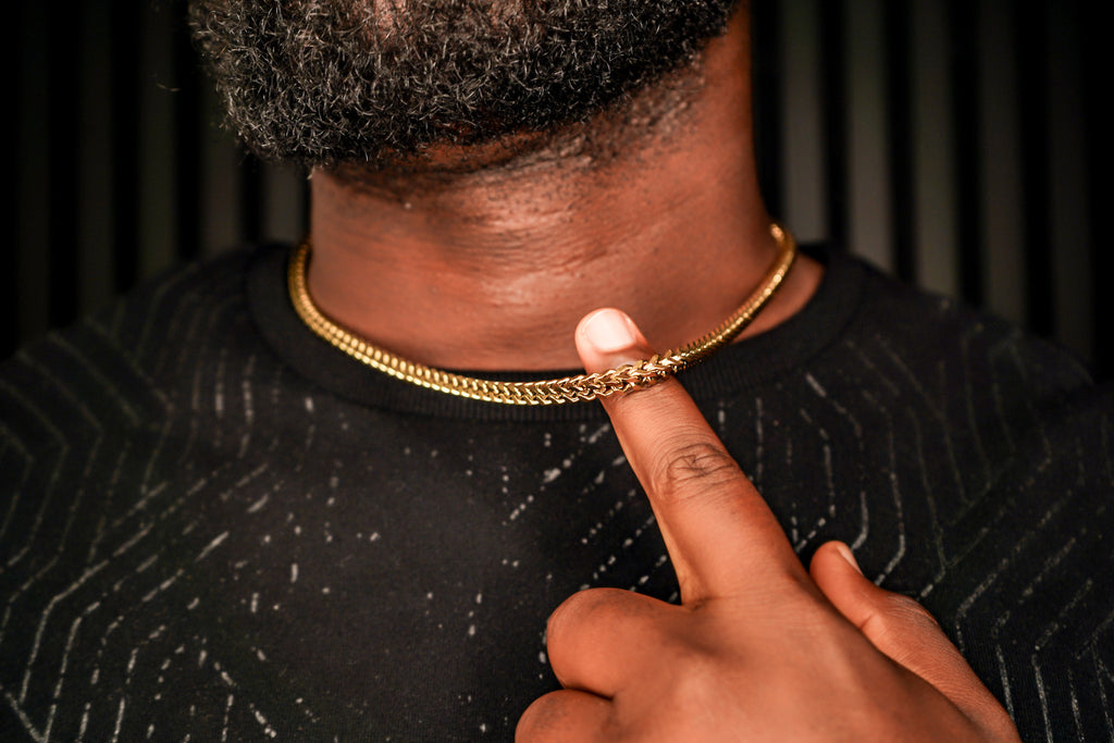 Person wearing a gold chain necklace with a finger pointing to it against a dark background -Signature-Franco-Chain-JCollins-Jewelry