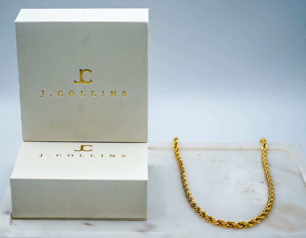 Gold chain necklace on a marble surface with J. Collins branded box in the background.
