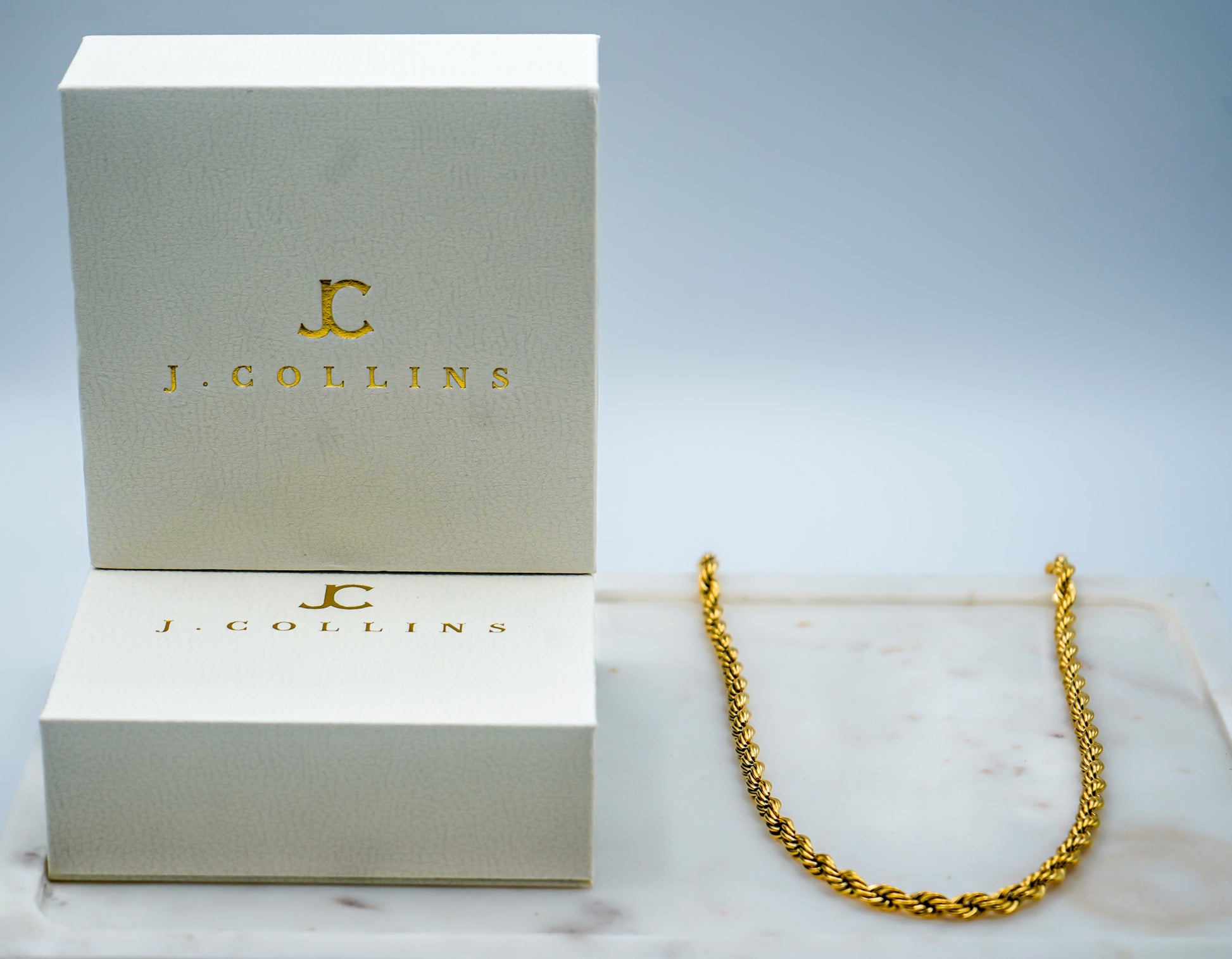Gold chain necklace on a marble surface with J. Collins branded box in the background.