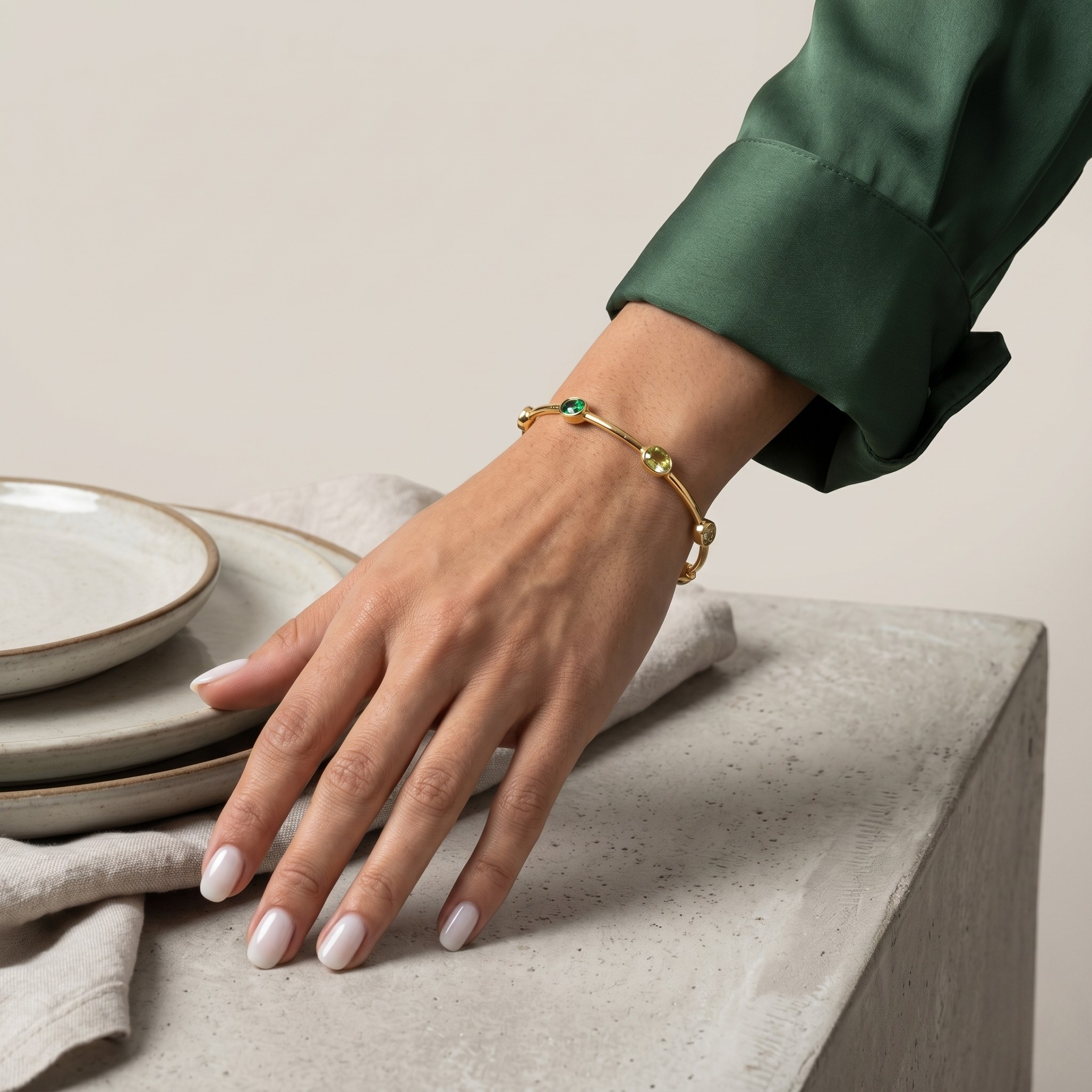 Model wearing a delicate gold chain bracelet with bezel-set round green gemstones and small pearl stations. Her hand is resting on a light grey textured surface next to stacked rustic ceramic plates. - Lush Inset Cuff - J. Collins Jewelry 