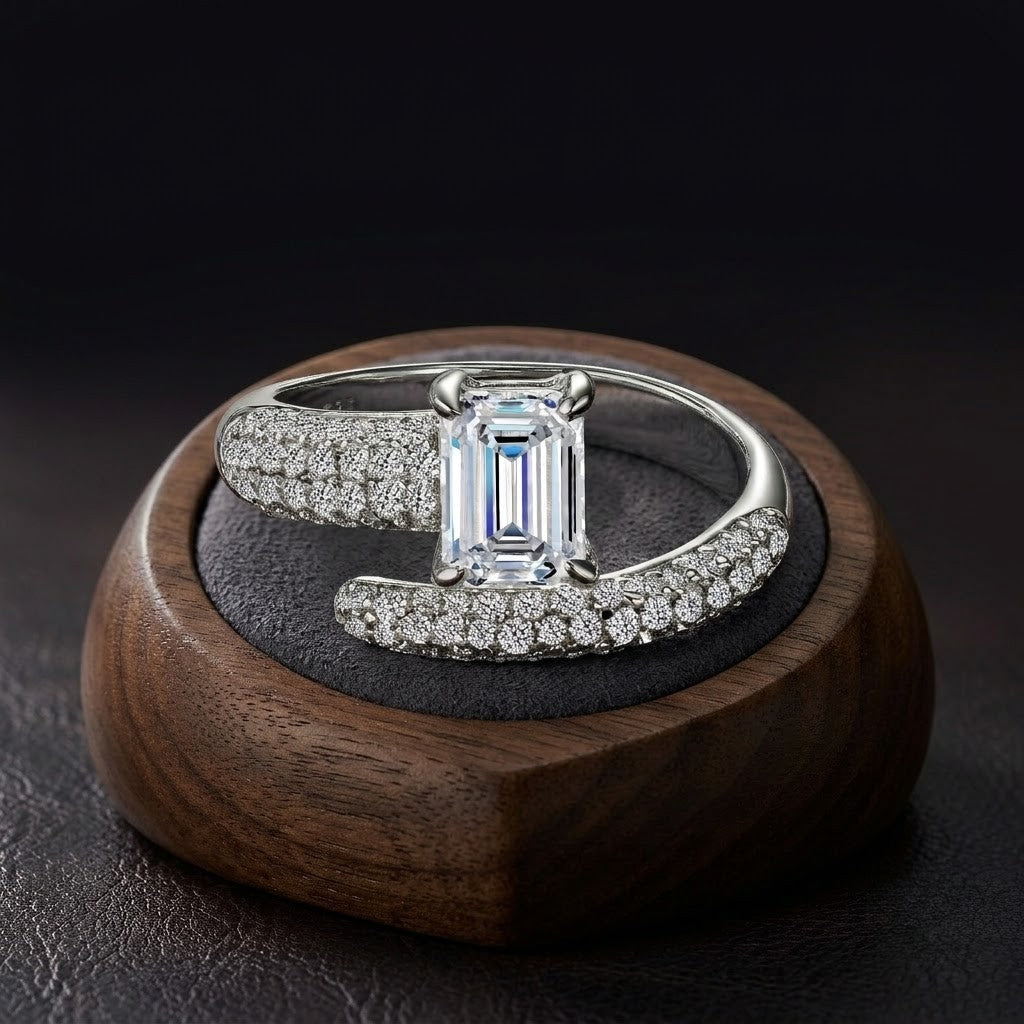 An emerald-cut diamond ring with pavé-set diamonds on a silver-toned band, displayed on a dark textured ring stand inside a round wooden box against a dark background - J.Collins Jewelry