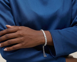 A close-up of a person's hand and wrist wearing a tennis bracelet with a row of clear stones and a dangling chain with small beads, set against a blue satin-sheen top- J.Collins Jewelry
