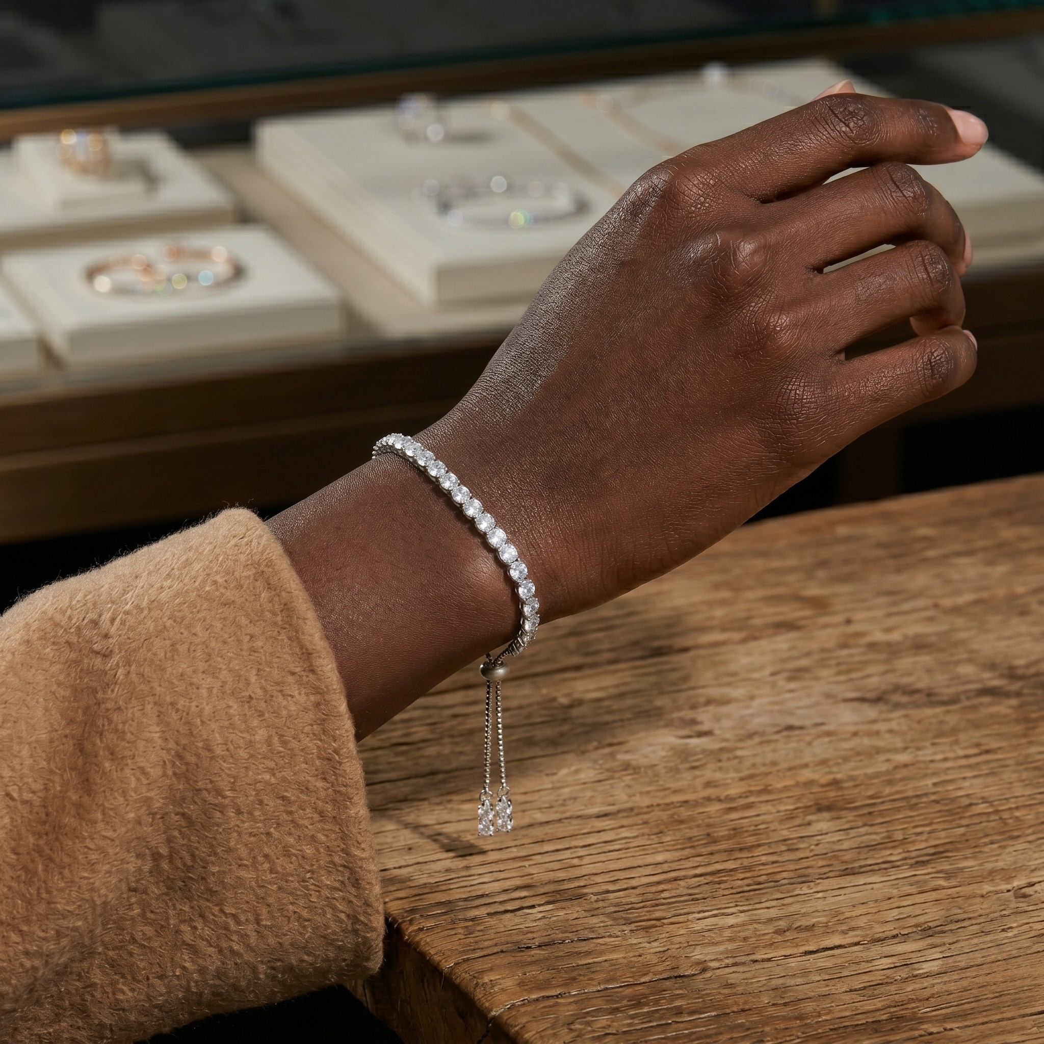 Hand wearing a diamond bracelet on a wooden surface with jewelry display in the background - Slider Tennis Bracelet - J. Collin’s Jewelry