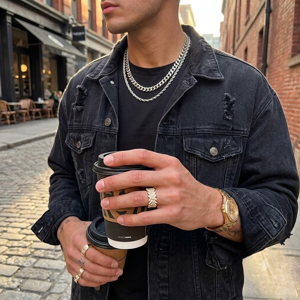 A man wearing a distressed black denim jacket, layered silver chain necklaces, and gold rings, holding a paper coffee cup on a cobblestone street - J.Collins Jewelry