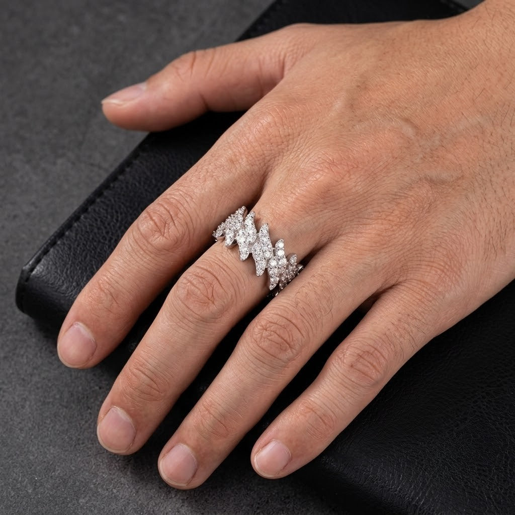 A close-up of a hand wearing a wide white gold ring set with multiple small clear stones in a wavy, layered design, resting on a black textured surface - J.Collins Jewelry