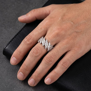 A close-up of a hand wearing a wide white gold ring set with multiple small clear stones in a wavy, layered design, resting on a black textured surface - J.Collins Jewelry