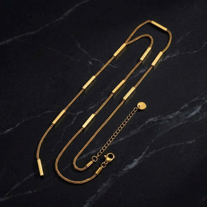 A gold-toned snake chain necklace with rectangular bar links, a lobster clasp, and an adjustable extension chain with a small round tag, displayed on a dark marble surface - J.Collins Jewelry