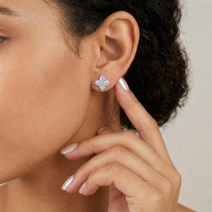 A close-up of a model's ear wearing a small silver-toned stud earring with a diamond-encrusted quatrefoil design, with her hand resting near her face against a soft light gray background.
