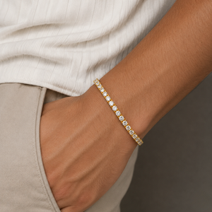 A close-up of a person's wrist wearing a delicate gold tennis bracelet with small clear stones, styled with a cream ribbed top and beige trousers - J.Collins Jewelry