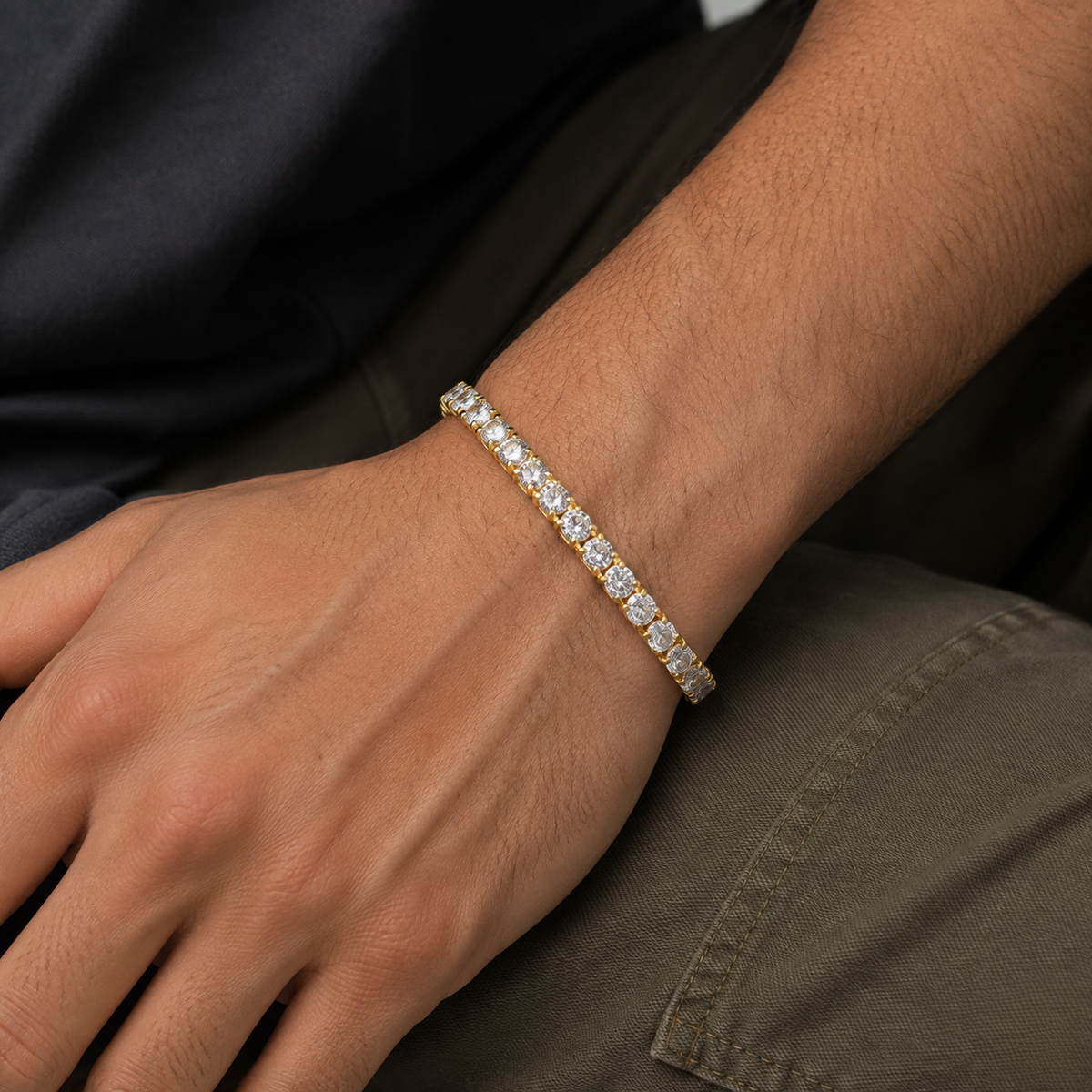 An upclose gold tennis bracelet on a model's hand positioned on gray jean cargo pant - J.Collins Jewelry