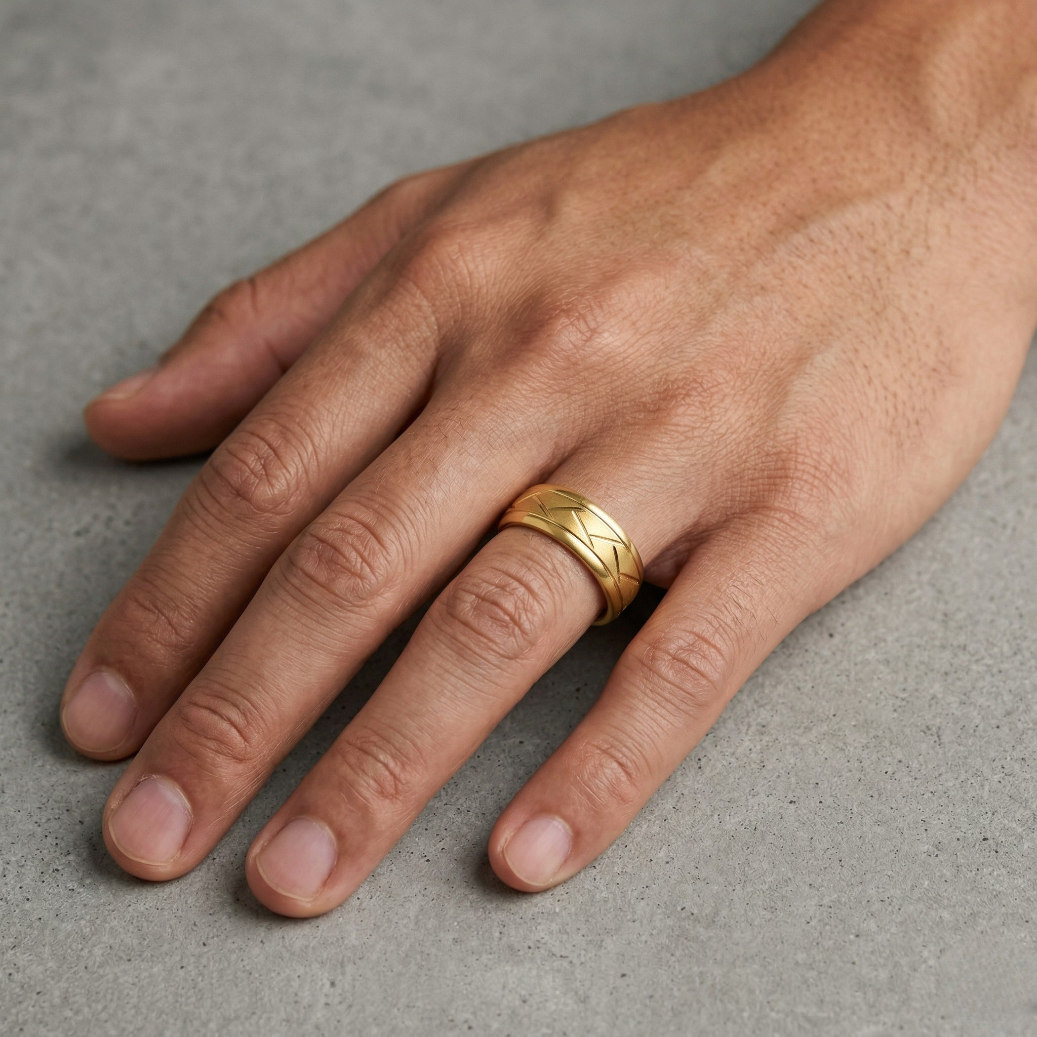 Man wearing wide gold band ring with engraved cross-hatch pattern on ring finger, men's textured wedding ring on concrete surface. J. Collins Jewelry 