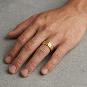 Man wearing wide gold band ring with engraved cross-hatch pattern on ring finger, men's textured wedding ring on concrete surface. J. Collins Jewelry 