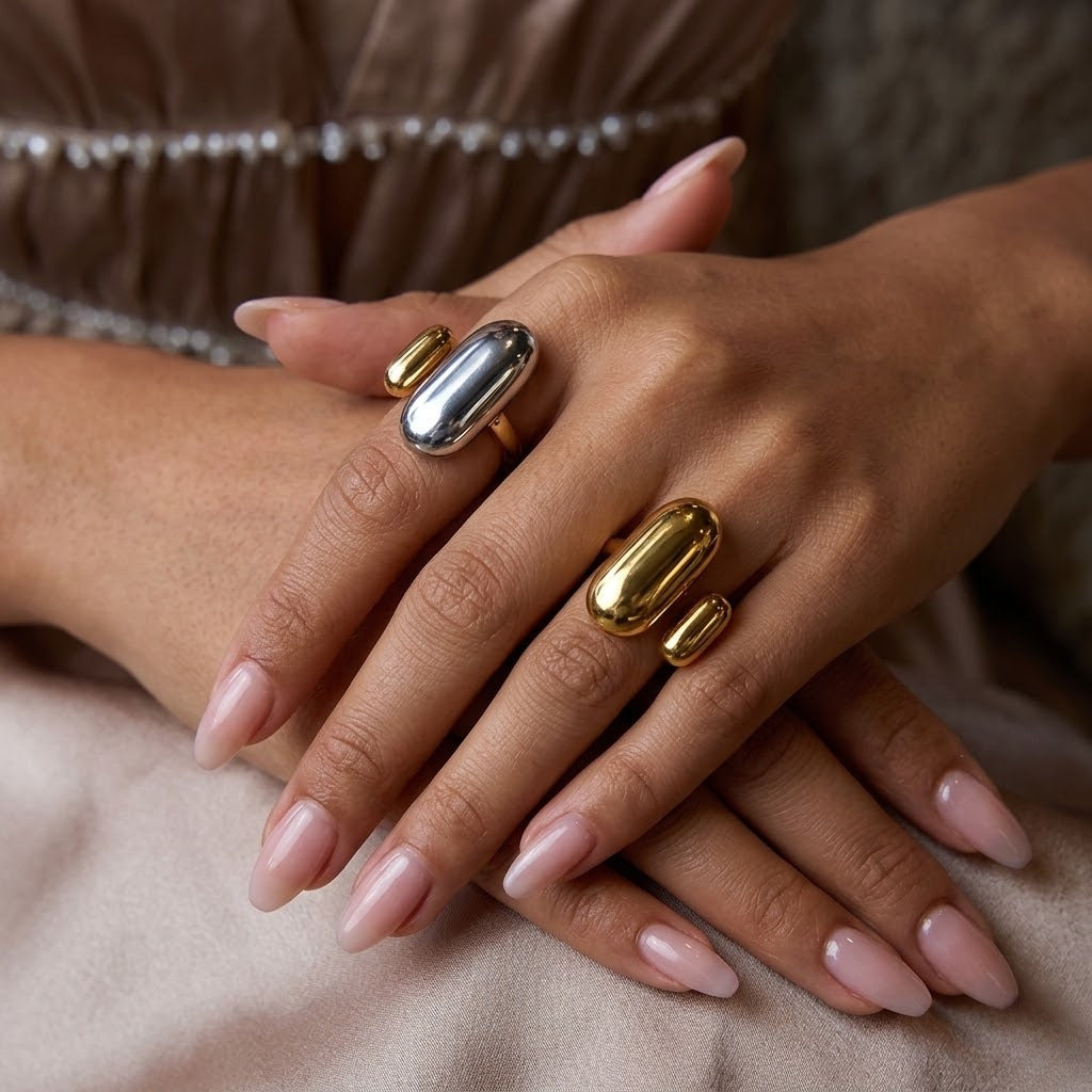 A close-up of a person's hands wearing two statement rings with smooth, elongated oval stones in polished gold and silver, set against a soft beige fabric background - J.XCollins Jewelry