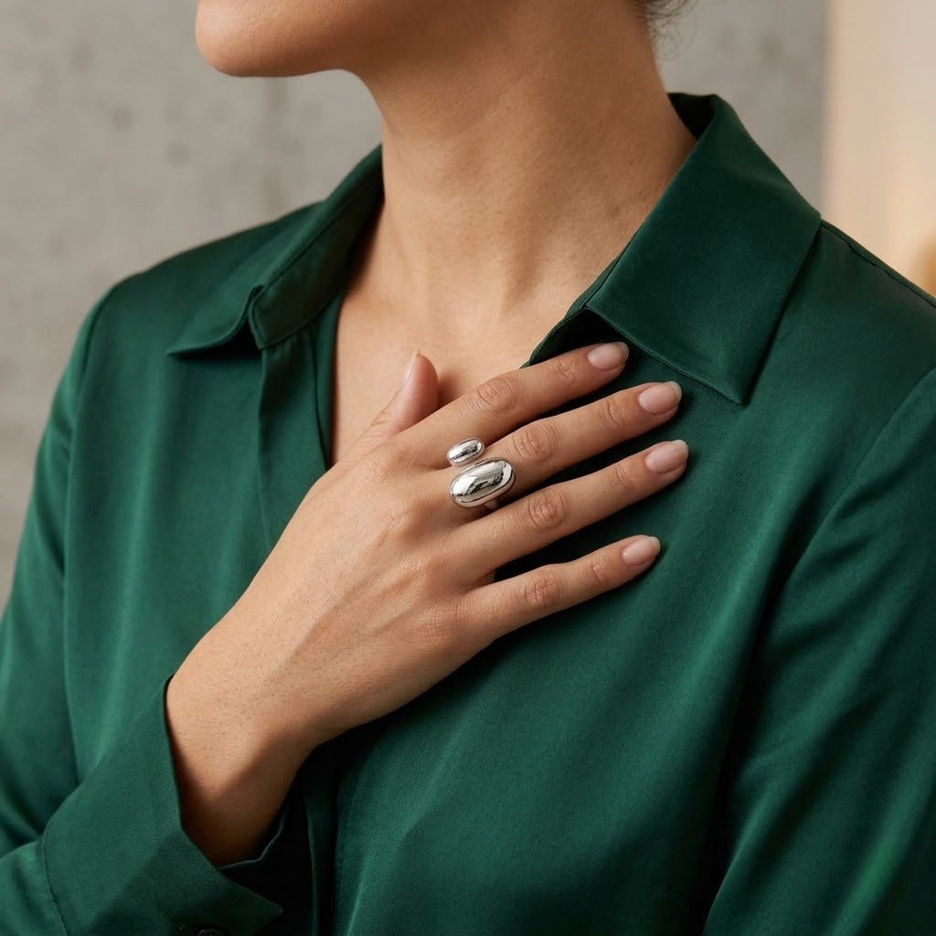 A close-up of a woman wearing a dark green satin shirt and a silver-toned double ring set on her fingers, with her hand resting on her chest - J.Collins Jewelry