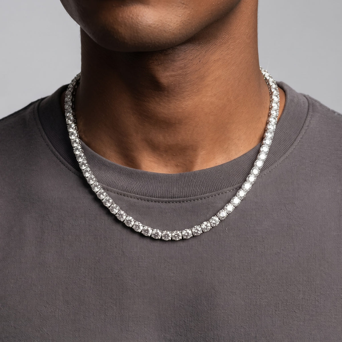 A close-up of a person wearing a silver-toned tennis necklace with clear stones, paired with a dark gray crewneck t-shirt against a neutral gray background - J.Collins Jewelry