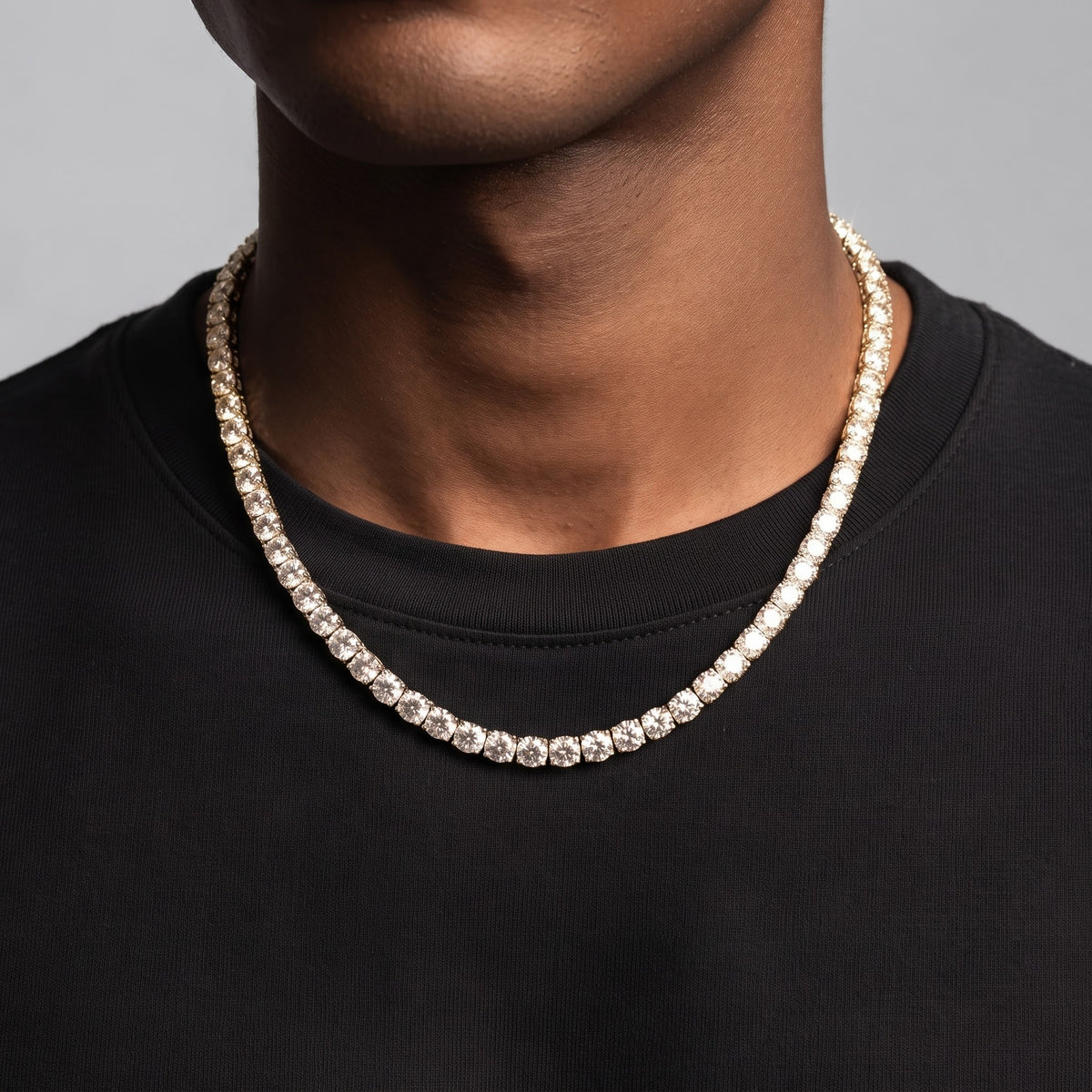 A close-up of a person wearing a silver-toned tennis necklace with clear stones, styled over a black crewneck shirt against a neutral gray background.