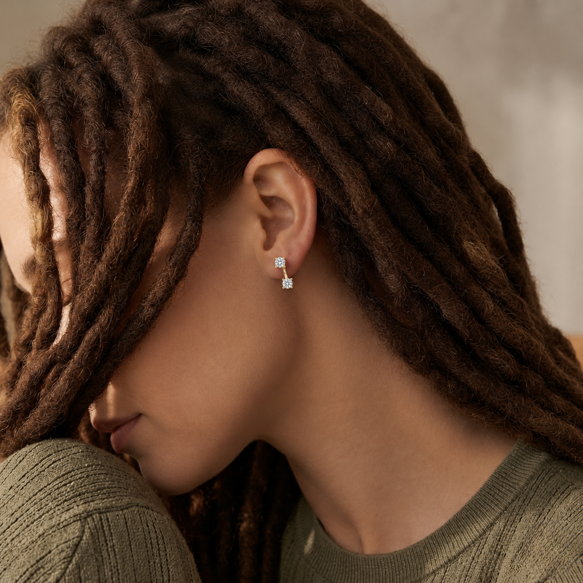 A close-up of a person with long dark brown dreadlocks wearing a small silver stud earring with a clear square gemstone. The person is wearing an olive green knit top against a softly blurred neutral background.