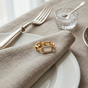 Gold chain link ring with diamond accents on a beige linen napkin, surrounded by silverware on a dining table.