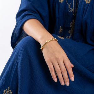 Gold cuff bracelet with pearl accents worn on a woman's wrist, paired with a blue embroidered dress.