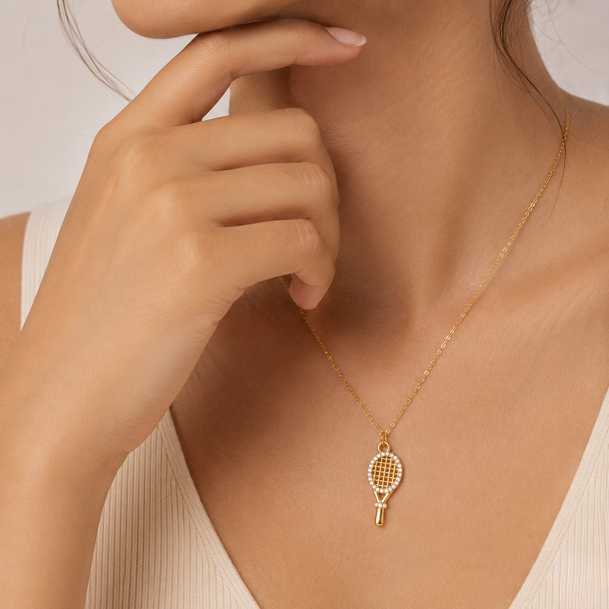 Tennis racket gold pendant necklace worn by a woman in a sleeveless top, hand near chin.
