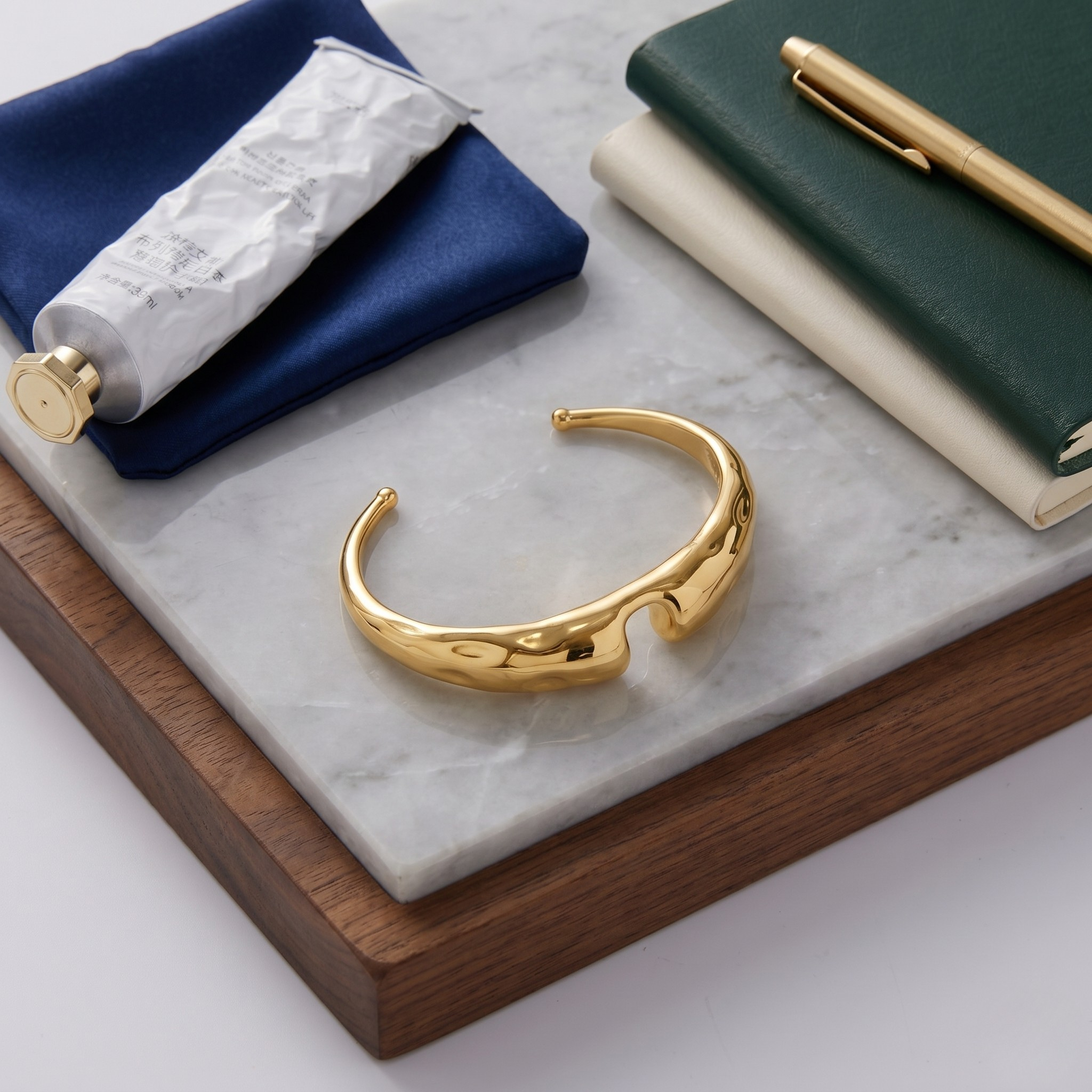 Gold bold sculpted cuff on marble and wood surface with notebooks, pen, and tube in lifestyle setting