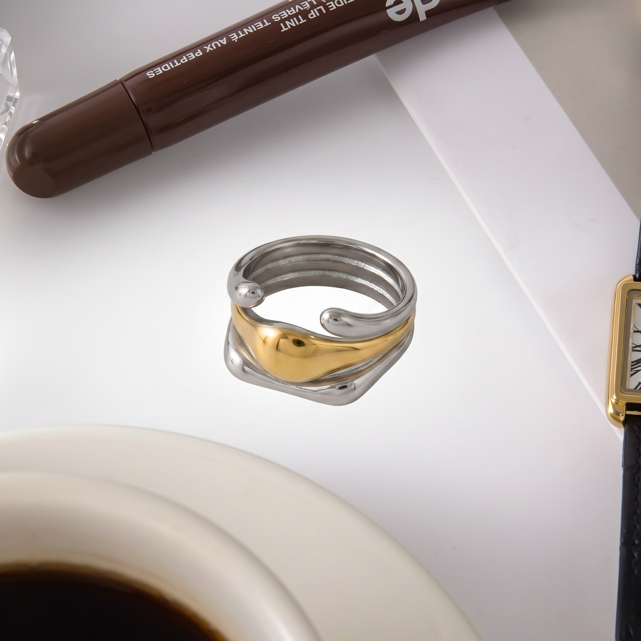 Two-tone interlocking metal band with gold and silver hues, placed near a watch on a light surface.