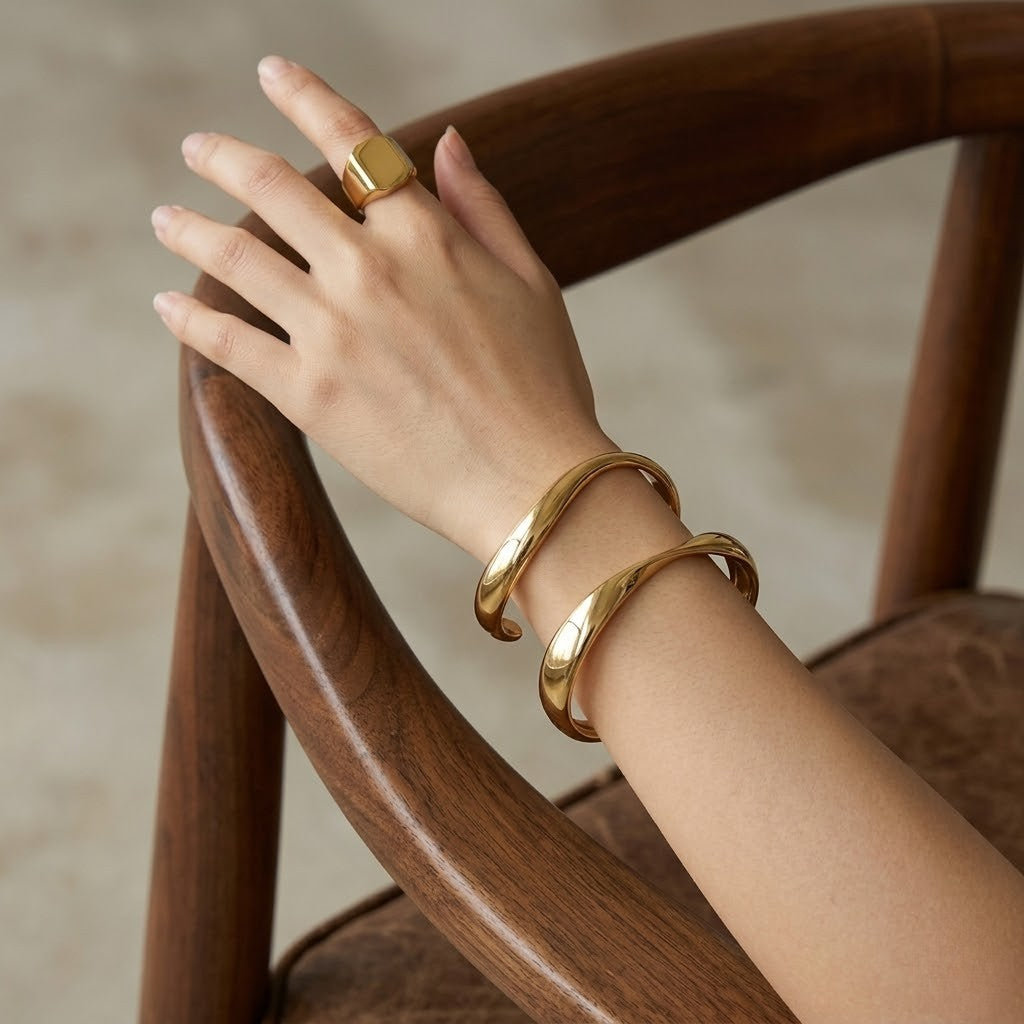 Gold Infinity Curve Bangles on wrist with matching gold signet ring, draped over wooden chair arm.