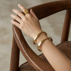 Gold Infinity Curve Bangles on wrist with matching gold signet ring, draped over wooden chair arm.