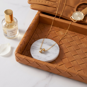Mirror Junction Charm Pendant gold necklace on leather tray with gloves, perfume, watch, and marble coaster.